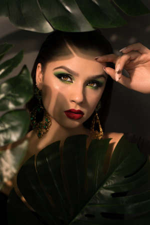 Close up face of beautiful young woman covering by green monstera green palm. Portrait of beauty...
