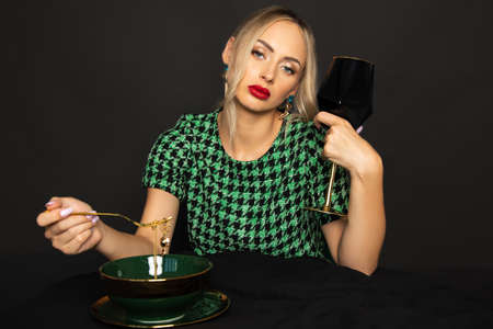 gorgeous blonde woman looks down on black background with glass of wine at table set with expensive dishes. Luxury lifestyle. Studio shot.の写真素材