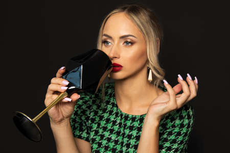 Luxurious young blonde woman in green dress on black background is tired, drinks alcohol from glass. concept of alcoholism in high society.の写真素材