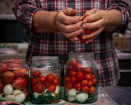 authentic photo of soiled female hands of elderly woman, cooking canned tomatoes from harvest in kitchen, putting vegetables in glass jars, cookingの写真素材