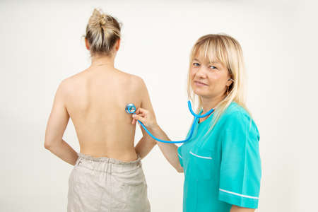 Attractive blonde doctor in turquoise medical uniform conducts medical examination with stethoscope....