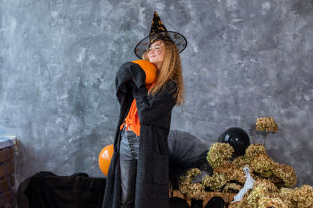 Teenage girl in hat among decor for Halloween holiday, huge pumpkin in hand. Humor photo. Black orange colorの写真素材