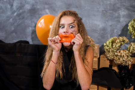 Teenage girl with shaggy hair inflates burst orange inflatable balloon among decor on halloweenの写真素材