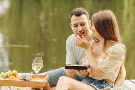 During picnic in nature, young woman feeds her man sweets from gift box. Sweet life. Happiness and serenity. Beautiful moments of life. Super day. Romantic relationships. Tenderness and care.の写真素材