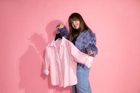 young woman fashion stylist holds hanger with shirt on pink background in studio. Basic wardrobe...
