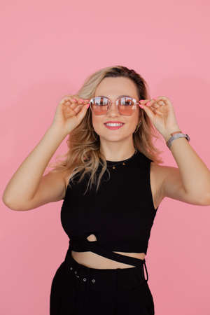 Portrait of charming blonde in sunglasses on pink background in studio. young woman smiles, looks happily into the frame. dressed in black dress with ribbons crossed on stomach.の写真素材