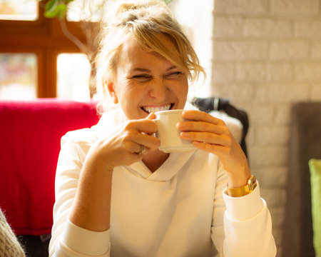 Photo of laughing cute young woman 40 years old drinking coffee, Beautiful curly hair woman sitting in cafe and enjoyingh coffee in hand in cup.の写真素材