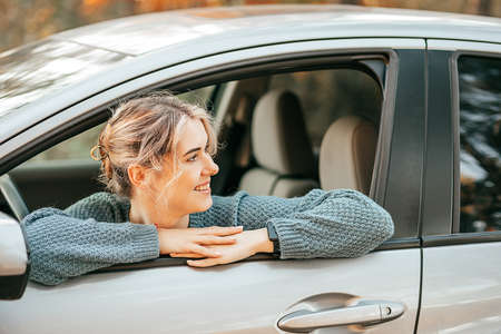 Photo of young beautiful blonde woman looked out of open car window, smiles cheerfully and looks around. Happy joyful journey, moment of relaxation, active lifestyle. High quality photoの写真素材