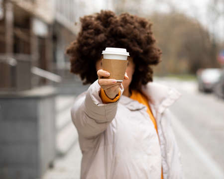 woman with fluffy curly dark hair hid behind disposable cup of tea or coffee with hot drink on city street. Shows it holding in hand. close-up photo, unrecognizable person.の写真素材