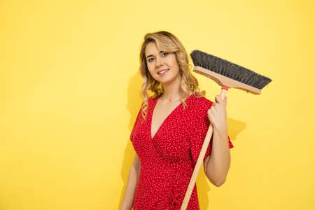 Happy young blonde woman in red dress on yellow background holds broom, smiling, cleaning service....