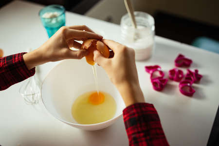 Hands of unrecognizable boy holding egg in his hands busy preparing dough for baking in kitchen at home. Hobby, cooking at home, over shoulder viewの写真素材