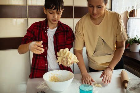 mom and son are cooking together in kitchen. boy is holding dough in hands, wearing plaid red shirt. preparing, cooking home kitchen at home. Relations with teenagers. Hobbies.の写真素材