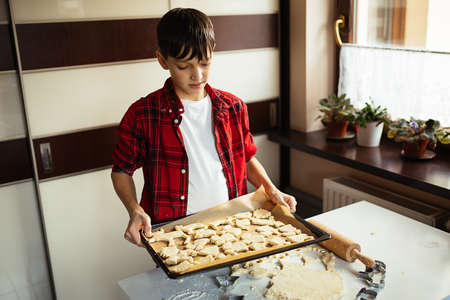 Boy teenager in checkered shirt in red, holds in his hands Baking sheet covered with parchment with cookies. Christmas cut-off biscuits. Animal shape. Heading to oven.の写真素材