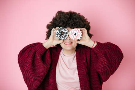 Close up portrait of excited caucasian woman afro hairstyle holding donuts against her eyes, hiding, fun with food, pink backgroundの写真素材
