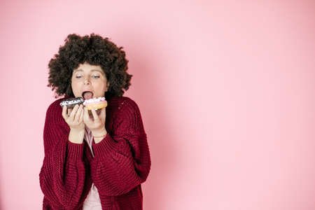 Young woman, afro hairstyle. Trying to eat 2 bottoms at once, concept addiction on food, pink background. Good appetite, unhealthy food.の写真素材