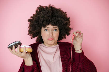 Close-up of shocked and upset beautiful afro hairstyle woman holding donut in hands and measuring tape. Concept of rejection of high-calorie foods and healthy lifestyle choice. addiction conceptの写真素材