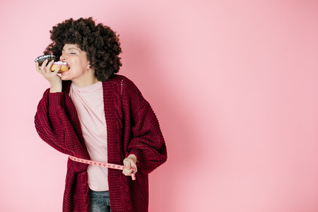 Woman on pink background bites off appetizing donut, hairstyle, African style. Holds it in her hands. Measurement tape. Humorous photography. Diet. Proper nutrition.の写真素材