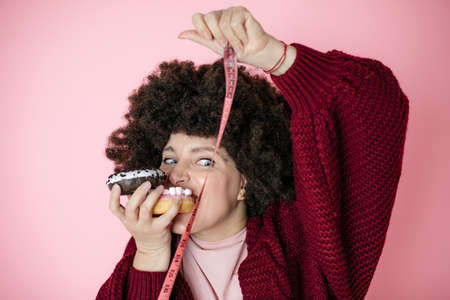 Woman holding donut in hand and check out his body fat with measuring tape. humor photo, fun, joy, dieting, addiction concept, pink background, afro hairstyleの写真素材