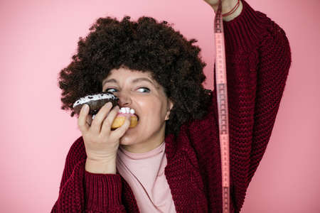 Woman holding donut in hand and check out his body fat with measuring tape. humor photo, fun, joy, dieting, addiction conceptの写真素材