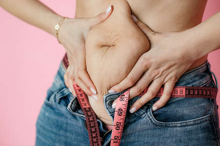 Measuring tape around waist, dieting concept, pink background, jeans, fat fold in hand, diastase,...