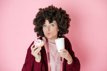Woman, afro hairstyle. Holds donat in hands And paper cup with hot drink, coffee or tea, looks into camera, measuring tape hangs around neck, choice of healthy food or Sweets. pink backgroundの写真素材
