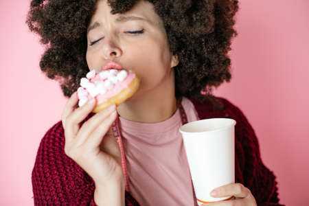 40 years woman Afro hairstyle enjoying donut and cup of coffee. measurement tape hangs around neck. Addiction to sweet foods. Unhealthy, diet. Pink backgroundの写真素材