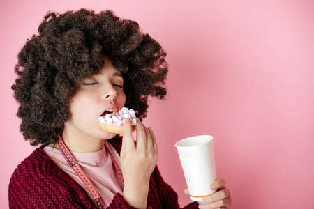 40 years woman Afro hairstyle enjoying donut and cup of coffee, measurement tape hangs around neck. Addiction to sweet foods. Unhealthy, diet. Pink backgroundの写真素材