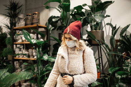 young blonde woman in knitted hat and faux fur coat chooses perennial plant in flower shop, flower...