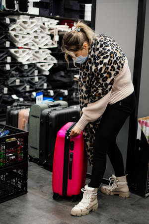 Woman in casual clothes, mask and leopard scarf is choosing bright pink suitcase in department of bags in supermarket. Tourism and travel. Trips to pandemic. Move. Shopping. Lifestyle.の写真素材