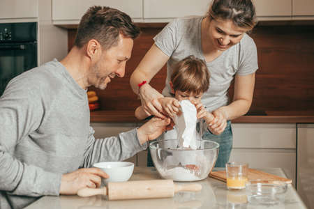 Happy young family with cute little preschooler kid boy 4-5 years old have fun baking cookies or pastry in modern kitchen together, mom and dad teach small children doing bakery cooking at homeの写真素材
