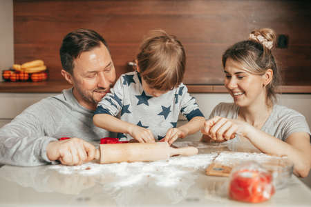 Happy family mother, father and son baking Christmas cookies on cozy kitchen at home, Little boy is helping mom and dad in making cookies. Spending time together, Use rolling pin to roll out dough.の写真素材