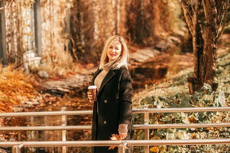 Illuminated by golden warm light, gorgeous blonde with cup of hot coffee in her hands stands in...