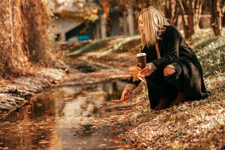 Golden landscape of autumn city park evokes nostalgic memories of sophisticated blonde in black cashmere coat leaning towardscolorful stream, she forgot about cup of coffee in her hand. lifestyles.の写真素材