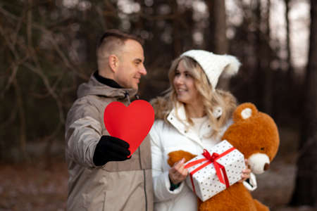 Symbol of Valentine's Day holiday is red heart in hands of cute man hugging his beloved woman, who has gift box and soft teddy bear in his hands. concept of dating. Lovers' Holiday. love emotions.の写真素材
