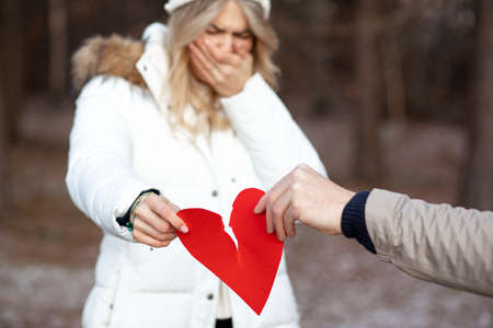 Woman is crying inconsolably, covering her face with her hand. Man's and woman's hand breaks paper red heart, symbol of their love. Concept of breaking up relationship. Abusive relationships.の写真素材