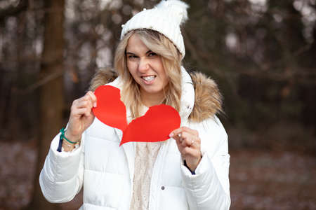 Blonde woman in winter jacket and knitted hat tears red heart in anger,symbol of Valentine's Day, holiday of lovers. Concept of breaking up relationship. Pain of betrayal. Abusive relationships.の写真素材