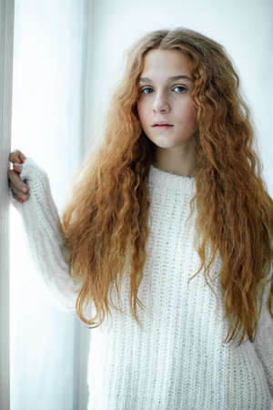 Blue-eyed teenage girl with luxurious long fluffy red hair stands near window in winter daylight and...