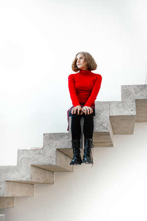 Teenage girl in red sweater has withdrawn into herself and looks away abstractedly, sitting on edge of steps of concrete staircase withou railing at very top. bullying. Misunderstanding.の写真素材