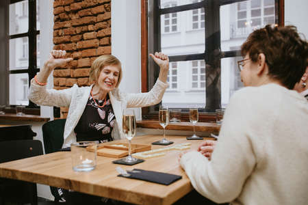 Smiling and laughing elderly woman with coral bracelet and chaplet in white jacket with her hands up playing dominoes in cafe with her friends at table with glasses of wine or champagne on itの写真素材