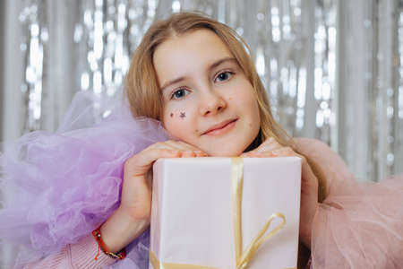 Charming fair-haired child girl having festival lilac and pink dress and stars on her face, holding white present box, sitting indoors possibly in cafe with decorated background of silvery spangleの写真素材