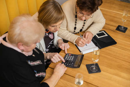Top view of intelligent good-looking elderly women meeting in modern cafe with orange walls , having calculator and notebook on table, dicussing prices, counting and writing down figuresの写真素材