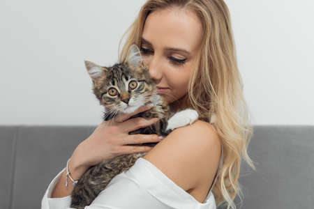 Portrait of young blond woman holding brown white and black cat looking at camera. Wearing bracelet. Gently hug kittenの写真素材
