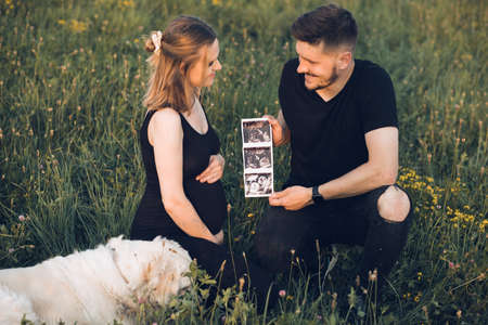 Young pregnant wife and husband walking at meadow, hold ultrasound photos. Couple waiting for baby,...