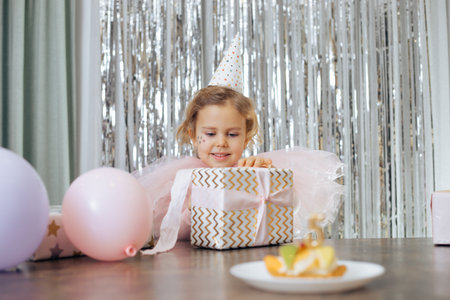 Cute little smiling girl with short curly hair and stars on face in pink poofy dress, cap unwrap...