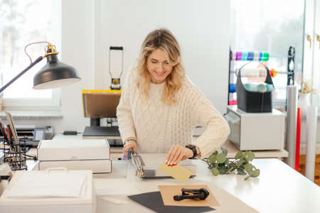 Professional smiling blond woman in sweater worker using printer cutter in workshop. Print office. Carving sheets paperの写真素材