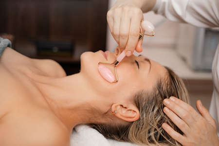 Attractive woman facial massage Gua Sha pink lifting stone roller. Close up. Beauty spa salon. Anti aging therapy. Careの写真素材