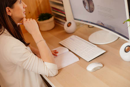 Young pretty working woman with long dark hair sitting at desk with notebook, white keyboard, mouse,...