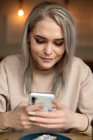 Vertical photo of upset tired woman in beige sweater, with manicure, cafe with plate of food, chatting online. Close upの写真素材