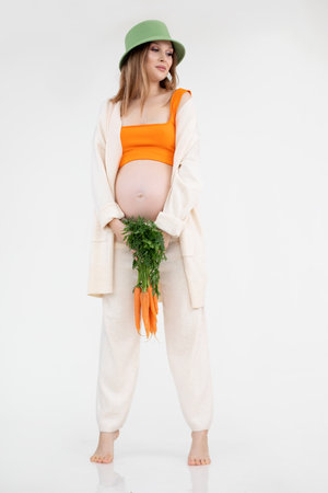Vertical photo of shy barefoot tiptoe smiling attractive woman, green hat staying, bunch of carrots.Pregnancy depilationの写真素材