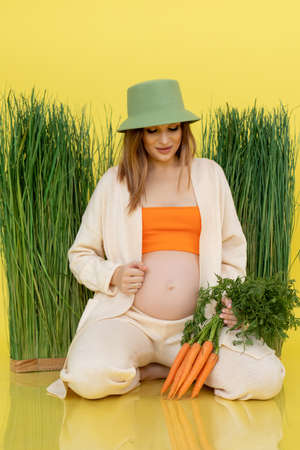 Vertical of glad satisfied pregnant woman sitting in green hat near grass, looking tummy with raw carrots. Yellow viewの写真素材